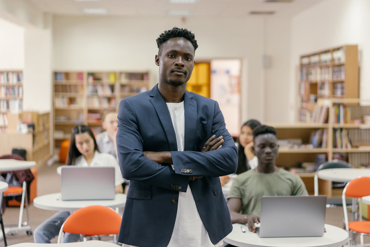 Teacher standing confidently in a library classroom with diverse students using laptops.